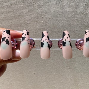 May include: Five long, matte, artificial nails with a cow print design. The nails are a light pink with black spots and a black tip. Each nail has a light blue heart shape near the base.