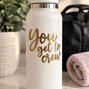 May include: A white insulated water bottle with a handle on the lid, featuring the gold text "You get to crew" printed on the front. The bottle has a smooth matte finish and a metallic silver band located just below the screw-top lid assembly.