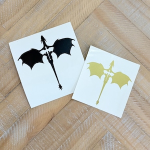 May include: Two square white stickers with dragon silhouettes. One dragon is black, the other is gold. The dragons have outstretched wings and long tails. The stickers are on a light brown wooden surface.