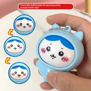 Op de afbeelding: Een lichtblauwe poppetjesvormige sleutelhanger met een veranderend gezicht. De pop heeft een blauwe capuchon, een wit gezicht en roze wangen. De tekst op de afbeelding luidt: Druk een seconde op de poppenknop om het gezicht te veranderen.