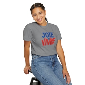 Puede incluir: Una camiseta gris de manga corta con el texto "JOIE" en letras azules sobre la palabra "de VIVRE" en letras rojas con estilo de pincelada. La camiseta tiene cuello redondo y se combina con unos pantalones vaqueros azules.