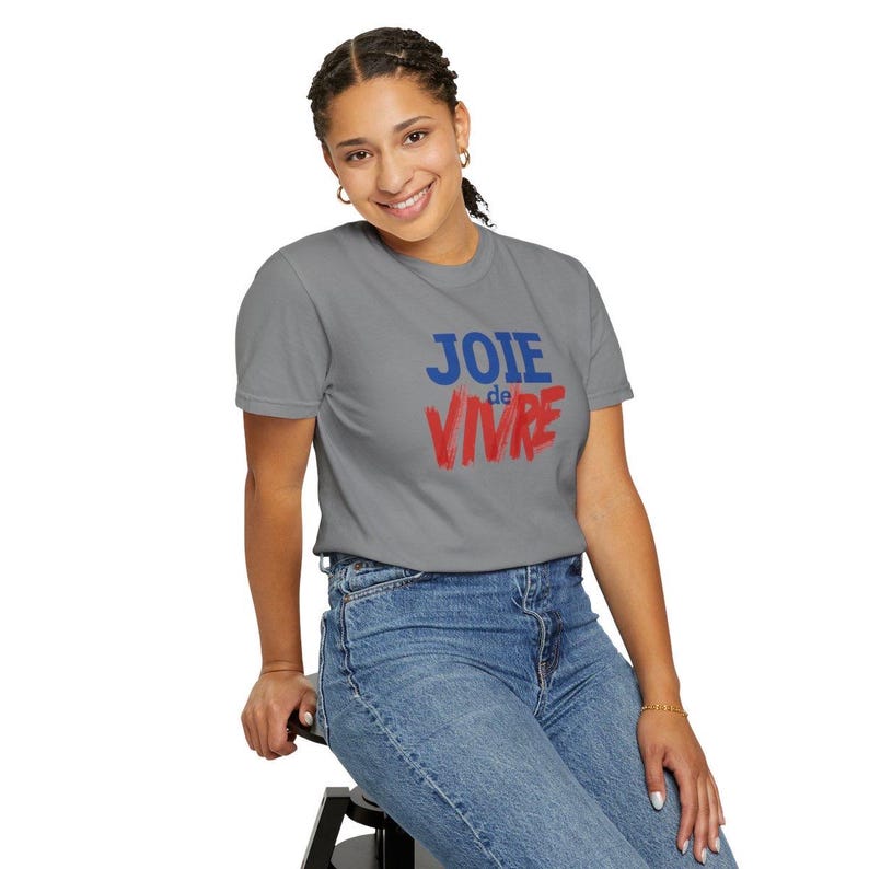 Puede incluir: Una camiseta gris de manga corta con el texto "JOIE" en letras azules sobre la palabra "de VIVRE" en letras rojas con estilo de pincelada. La camiseta tiene cuello redondo y se combina con unos pantalones vaqueros azules.