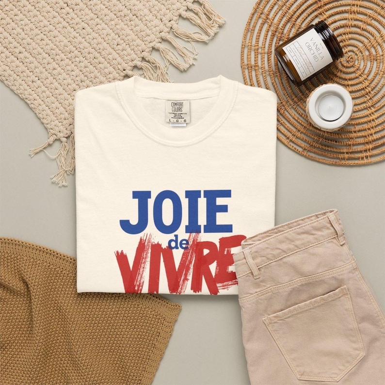 Puede incluir: Una camiseta de manga corta de color crema con el texto JOIE de VIVRE impreso en la parte delantera. La palabra JOIE es azul, la palabra de est&aacute; en letras azules peque&ntilde;as y la palabra VIVRE est&aacute; en letras rojas grandes y negritas.
