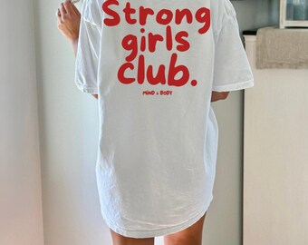 May include: A white short-sleeved t-shirt featuring the red text Strong girls club. printed in a bold, rounded font on the back. Below the main text, the words MIND & BODY are printed in a smaller, matching red font.