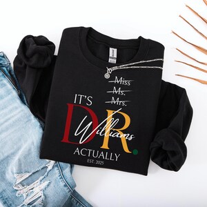 May include: Black crewneck sweatshirt with the text "IT'S DR. Williams ACTUALLY EST. 2025" in white, red, and gold lettering. The design includes crossed-out titles: Miss, Ms., and Mrs. A silver necklace is draped across the sweatshirt.