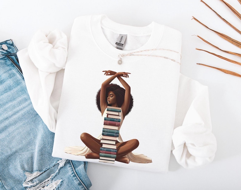 May include: White t-shirt with a graphic of a Black woman sitting with a stack of books forming a dress. The woman has her arms raised above her head and is looking to the side.