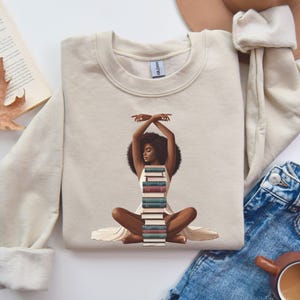 May include: A beige sweatshirt with a graphic of a Black woman sitting with a stack of books in her lap. The woman is wearing a white dress and has her arms raised above her head.