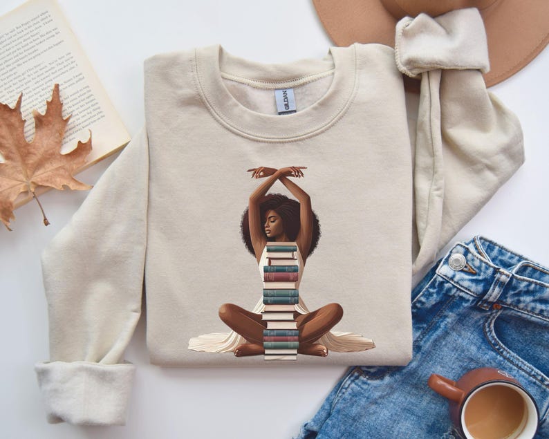 May include: A beige sweatshirt with a graphic of a Black woman sitting with a stack of books in her lap. The woman is wearing a white dress and has her arms raised above her head.