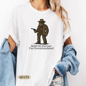 Może przedstawiać: Biała koszulka z postacią z kresk&oacute;wki Bigfoot w brązowym garniturze i kapeluszu, trzymającą pistolet. Tekst "Hold On Partner I'm Overstimulated" jest wydrukowany pod grafiką. Koszulka ma okrągły dekolt i kr&oacute;tkie rękawy.