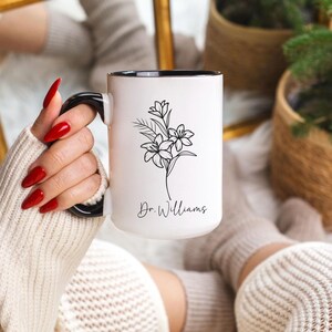May include: White ceramic mug with a black interior and handle. The mug features a floral line drawing and the name "Dr. Williams" in script. The mug is held by a person with red nail polish.