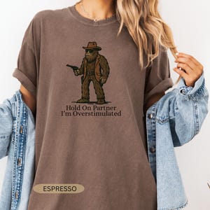 Może przedstawiać: Koszulka w kolorze espresso z postacią z kresk&oacute;wki Bigfoot w kapeluszu kowbojskim i kamizelce, trzymającą pistolet. Tekst "Hold On Partner I'm Overstimulated" znajduje się poniżej. Koszulka ma podwinięte rękawy.