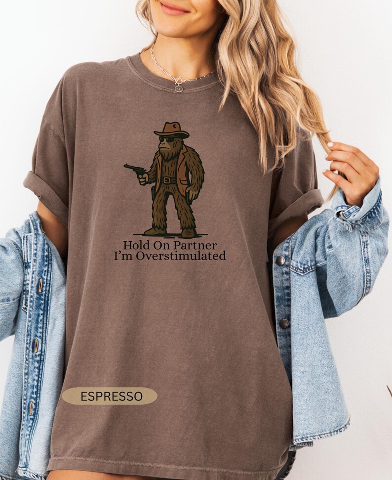 Może przedstawiać: Koszulka w kolorze espresso z postacią z kresk&oacute;wki Bigfoot w kapeluszu kowbojskim i kamizelce, trzymającą pistolet. Tekst "Hold On Partner I'm Overstimulated" znajduje się poniżej. Koszulka ma podwinięte rękawy.