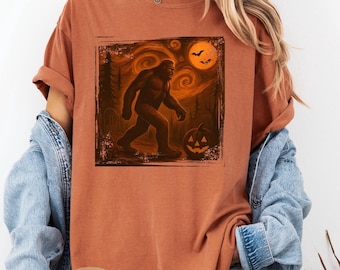 Może przedstawiać: Rdzawa koszulka z grafiką o tematyce Halloween. Grafika przedstawia postać Bigfoota idącą w lesie z pełnią księżyca, nietoperzami i dynią. Słowo "Yam" jest wydrukowane na dole.
