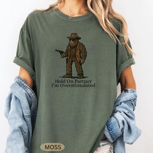 Może przedstawiać: Zielona koszulka w kolorze mchu z postacią z kresk&oacute;wki Bigfoot w kapeluszu kowbojskim i kurtce, trzymającą pistolet. Tekst "Hold On Partner I'm Overstimulated" jest wydrukowany poniżej. Koszulka ma luźny kr&oacute;j.