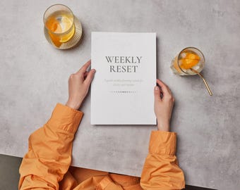 May include: A person holding a white planner with the words "WEEKLY RESET" on the cover. The planner is surrounded by two glasses of orange-infused water and an orange-colored long-sleeved shirt.