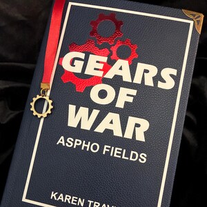 Puede incluir: Un libro azul marino titulado "GEARS OF WAR: ASPHO FIELDS" de Karen Traviss. La portada presenta gráficos de engranajes rojos y texto blanco. Se adjunta un marcapáginas de cinta roja con un dije de engranaje dorado. Protectores de esquina dorados añaden detalles.