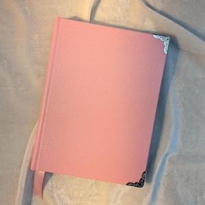 May include: A rectangular notebook with a textured light pink cover. The front cover features decorative silver-toned metal corner protectors with an intricate filigree pattern. A matching light pink ribbon bookmark extends from the bottom edge.