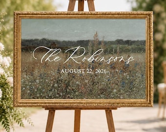 Puede incluir: Un letrero rectangular en un marco dorado ornamentado sobre un caballete de madera. El letrero presenta una pintura de paisaje de un campo con flores silvestres y el texto blanco The Robinsons AUGUST 22, 2026 centrado en la superficie.