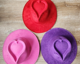 May include: Three fedora hats in red, pink, and purple. Each hat features a heart-shaped crown and a wide brim. The hats are made of a soft, suede-like material and are arranged on a light brown wooden surface.
