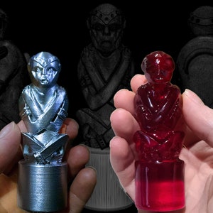 May include: Two small statues of a seated figure with arms crossed. One statue is made of silver metal and the other is made of red glass.