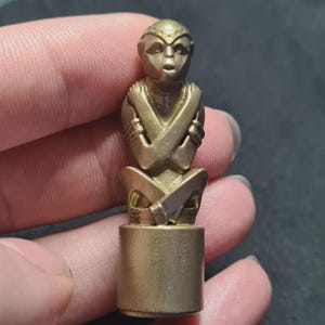 May include: A small, brass-coloured figurine of a seated monkey with its arms crossed. The monkey has a detailed face and is mounted on a cylindrical base. The figurine is made of a metallic material and has a polished finish.