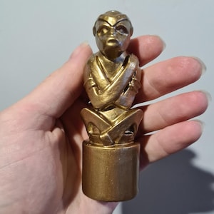 May include: A gold-colored figurine of a person with their arms crossed, sitting on a round base. The figurine is made of a smooth, shiny material.