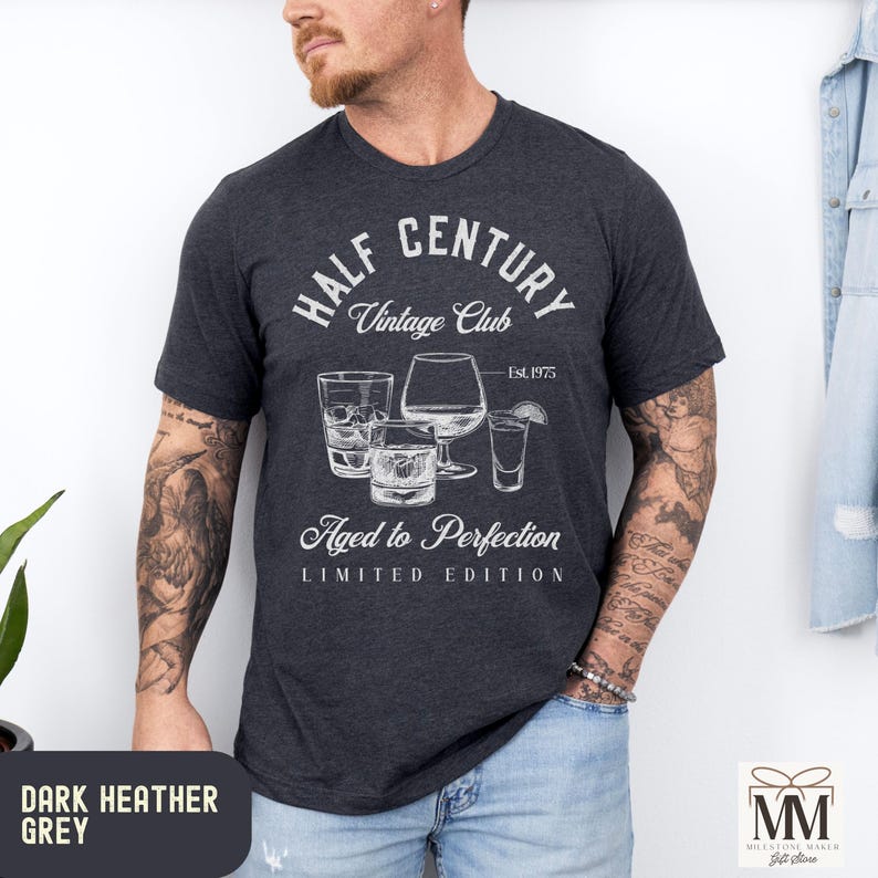 May include: Dark heather grey t-shirt with a white graphic design. The design includes the text "HALF CENTURY Vintage Club" and "Aged to Perfection". The graphic also features illustrations of various glasses. The shirt is a limited edition.
