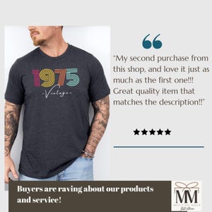 以下が含まれることがあります： レインボーカラーで「1975」とプリントされたダークグレーのTシャツ。下に「ヴィンテージ」の文字が書かれている。腕にタトゥーを入れた人がTシャツを着ている。