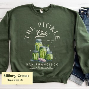 Puede incluir: Sudadera verde militar con un dise&ntilde;o de "The Pickle Club" y tarros de pepinillos. El dise&ntilde;o incluye el texto "San Francisco Gourmet Pickles and Brines" y "Est. 1892".