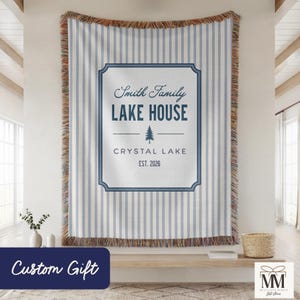 Peut inclure: Une couverture tissée personnalisée avec des rayures verticales bleues et blanches et une bordure à franges. Le centre présente un motif encadré avec le texte "Smith Family LAKE HOUSE CRYSTAL LAKE EST. 2026". Une bannière bleue en bas indique "Custom Gift".