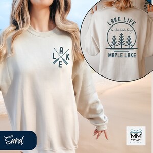 K&ouml;nnte beinhalten: Ein beiger Pullover mit einer Grafik aus gekreuzten Rudern und dem Wort "LAKE" auf der Vorderseite. Die R&uuml;ckseite des Pullovers zeigt eine Grafik mit B&auml;umen und dem Text "LAKE LIFE is the best life MAPLE LAKE".