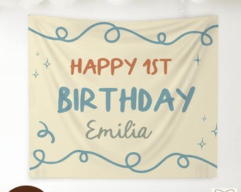 May include: A beige fabric banner with the text "HAPPY 1ST BIRTHDAY Emilia" in blue and orange lettering. The banner has blue swirls and star accents, suitable for a first birthday celebration.