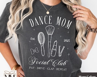 以下が含まれることがあります： 白い文字で「Dance Mom Social Club」と書かれた濃いグレーのTシャツ。テキストは、メイクブラシ、ヘアブラシ、口紅、ヘアピンを白く縁取ったもので囲まれています。「Est. 2025」というテキストは、「Dance Mom」というテキストの下にあります。