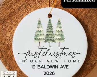 May include: A round white ceramic ornament featuring three watercolor-style green pine trees. The text reads: first christmas IN OUR NEW HOME 19 BALDWIN AVE 2026. A gold string is attached through a hole at the top for hanging the holiday decoration.