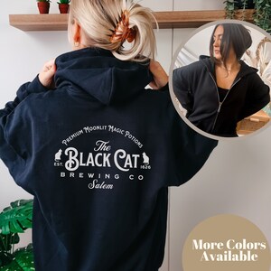 May include: Black hooded sweatshirt with a white graphic print of a black cat logo. The logo says "The Black Cat Brewing Co. Salem" and "Premium Moonlit Magic Potions" around the logo. The text "Est. 1626" is on the right side of the logo.
