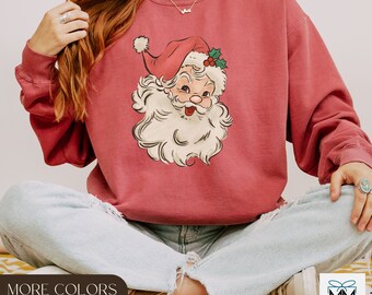 May include: A red sweatshirt with a vintage illustration of Santa Claus's face. The sweatshirt has a crew neck and long sleeves. The text "MORE COLORS AVAILABLE" is visible in the bottom left corner of the image.
