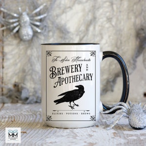 May include: White ceramic mug with a black rim and handle. The mug features a black and white graphic of a raven and text that reads "The Salem Massachusetts Brewery and Apothecary Elixirs - Potions - Brews".