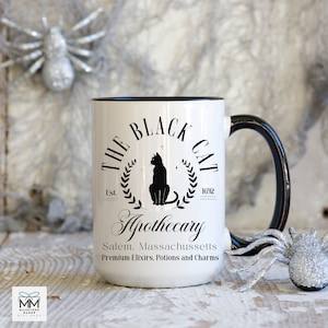 May include: White ceramic mug with a black rim and handle. The mug features a black and white illustration of a black cat sitting in front of a wreath with the text "The Black Cat Apothecary Est. 1692 Salem, Massachusetts Premium Elixirs, Potions and Charms".