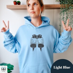 Op de afbeelding: Een lichtblauwe hoodie met een zwarte afbeelding van een paar schaatsen. De hoodie heeft een capuchon met trekkoord en een kangoeroezak.