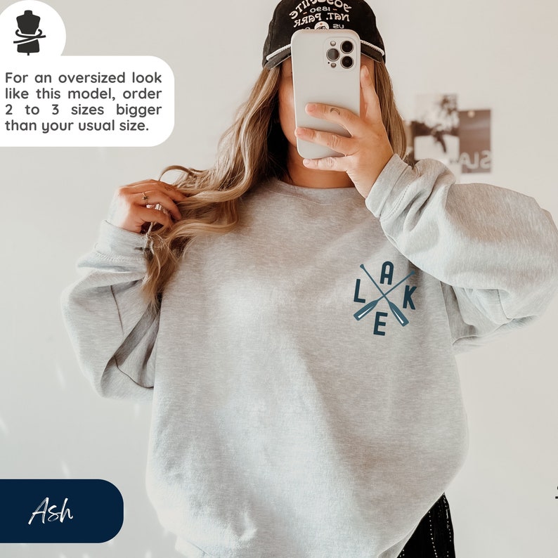 K&ouml;nnte beinhalten: Ein graues Sweatshirt mit einer blauen Grafik aus gekreuzten Rudern und dem Wort "LAKE" auf der Vorderseite. Der Text "Ash" ist in der linken unteren Ecke des Bildes sichtbar.