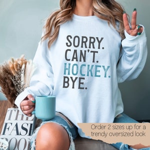 May include: A light blue sweatshirt with the text "SORRY. CAN'T. HOCKEY. BYE." in black and blue lettering. The text is in a distressed font. The sweatshirt is being worn by a person sitting on a couch. The text "Order 2 sizes up for a trendy oversized look" is visible in the bottom right corner of the image.