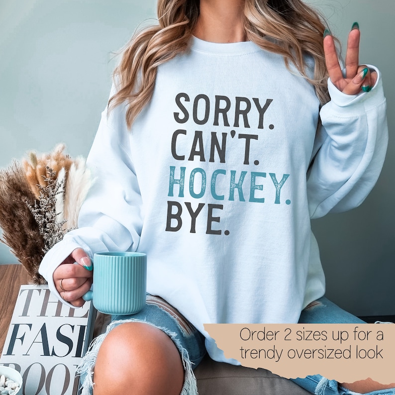 May include: A light blue sweatshirt with the text "SORRY. CAN'T. HOCKEY. BYE." in black and blue lettering. The text is in a distressed font. The sweatshirt is being worn by a person sitting on a couch. The text "Order 2 sizes up for a trendy oversized look" is visible in the bottom right corner of the image.