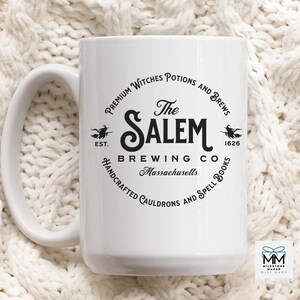Puede incluir: Taza de cer&aacute;mica blanca con un dise&ntilde;o gr&aacute;fico negro de un logotipo para "The Salem Brewing Co." El logotipo presenta una bruja en una escoba, el texto "EST. 1626" y las palabras "Premium Witches Potions and Brews", "Handcrafted Cauldrons and Spell Books" y "Massachusetts".