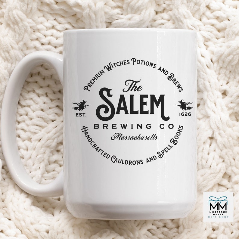 Puede incluir: Taza de cer&aacute;mica blanca con un dise&ntilde;o gr&aacute;fico negro de un logotipo para "The Salem Brewing Co." El logotipo presenta una bruja en una escoba, el texto "EST. 1626" y las palabras "Premium Witches Potions and Brews", "Handcrafted Cauldrons and Spell Books" y "Massachusetts".