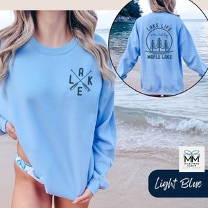 K&ouml;nnte beinhalten: Hellblauer Pullover mit einem Aufdruck von gekreuzten Paddeln und dem Wort "LAKE" auf der Vorderseite. Die R&uuml;ckseite des Pullovers hat einen Aufdruck von B&auml;umen und dem Text "LAKE LIFE is the best life MAPLE LAKE".