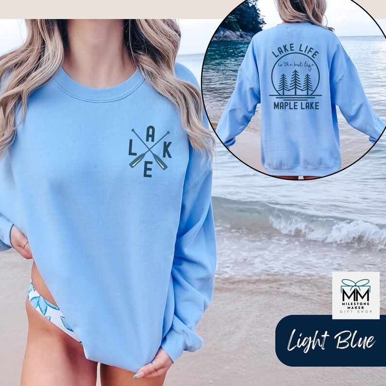 K&ouml;nnte beinhalten: Hellblauer Pullover mit einem Aufdruck von gekreuzten Paddeln und dem Wort "LAKE" auf der Vorderseite. Die R&uuml;ckseite des Pullovers hat einen Aufdruck von B&auml;umen und dem Text "LAKE LIFE is the best life MAPLE LAKE".
