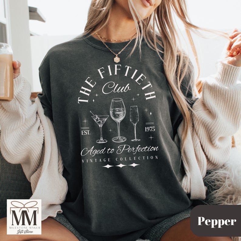 May include: Pepper-colored graphic tee with the text "THE FIFTIETH Club" and "Aged to Perfection". The design includes illustrations of three cocktail glasses. The shirt is from the "VINTAGE COLLECTION" and has the year "1975" printed on it.