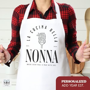 以下が含まれることがあります： 「La Cucina Della Nonna Est. 2024 Where Every Meal Is Made With Love」という黒い文字が書かれた白いエプロン。エプロンには、フォークとパスタの白黒のイラストが描かれている。