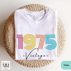 以下が含まれることがあります： 白いTシャツに、カラフルな1975年のグラフィックと「ヴィンテージ」という文字がプリントされています。