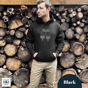 Op de afbeelding: Zwart hoodie met een afbeelding van een paar schaatsen. De hoodie wordt gedragen door een persoon die voor een muur van gestapelde boomstammen staat.